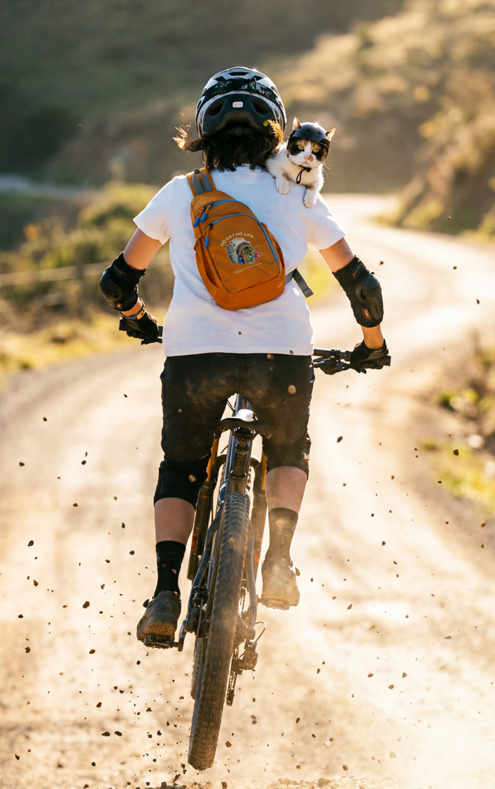 マウンテンバイクで山道を駆け抜ける人物・オレンジのNEKONI SLINGバッグ・肩に猫